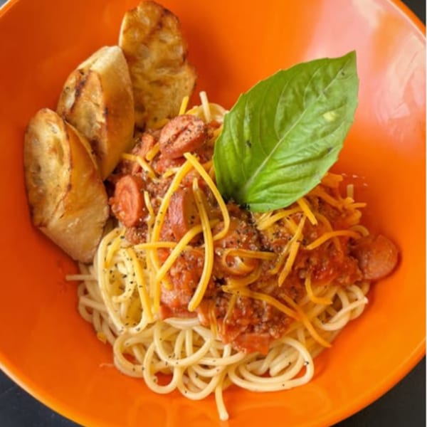 Filipino-style spaghetti with hot dogs, cheese, and basil in an orange bowl with garlic bread.