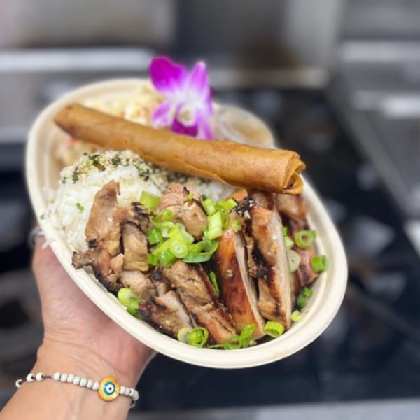 Hand holding a Filipino-Hawaiian OverRice bowl with grilled chicken, rice, lumpia, and a purple orchid.